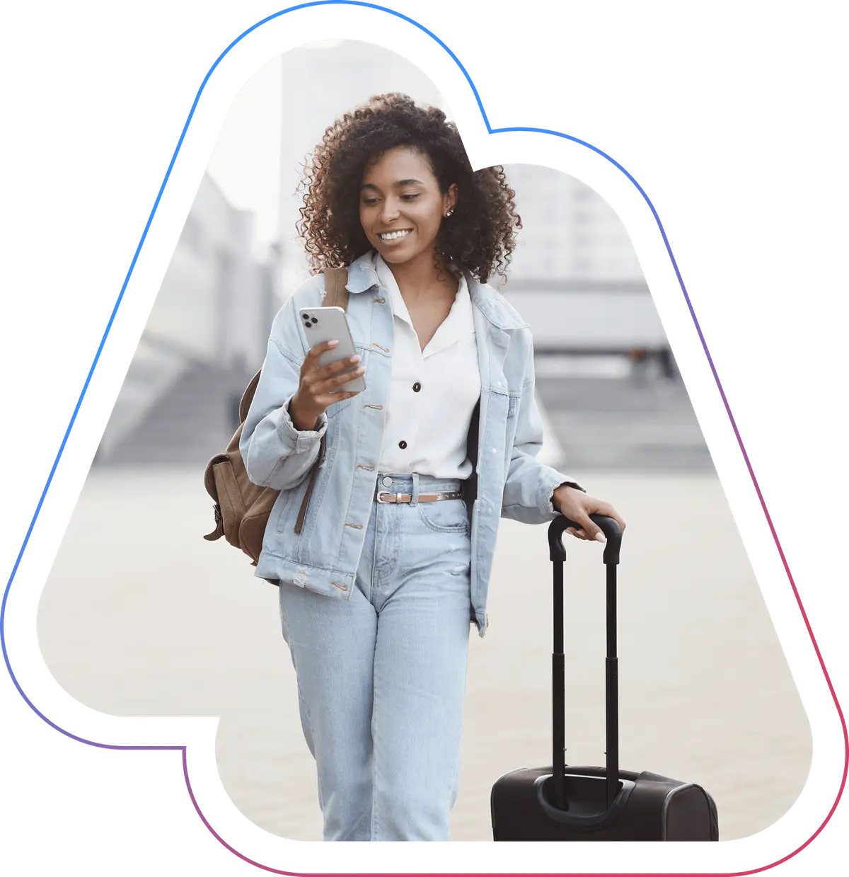 Smiling woman with curly hair in denim outfit, checking her phone while pulling a suitcase outdoors.